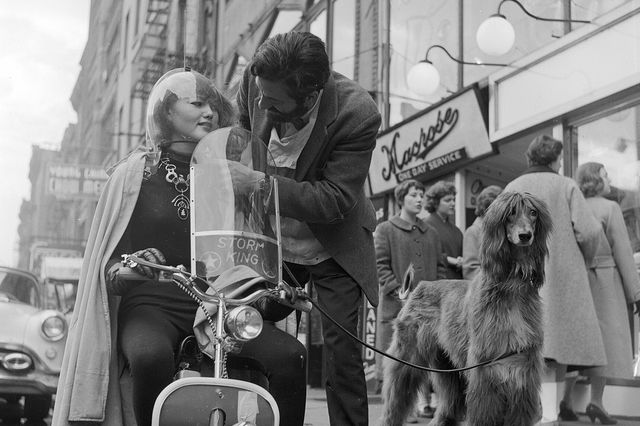 "American jeweller, Sam Kramer helping one of his 'Space Girls' with her motorbike helmet on a Greenwich Village street. His clipped Afghan Hound waits for him patiently. 1955."